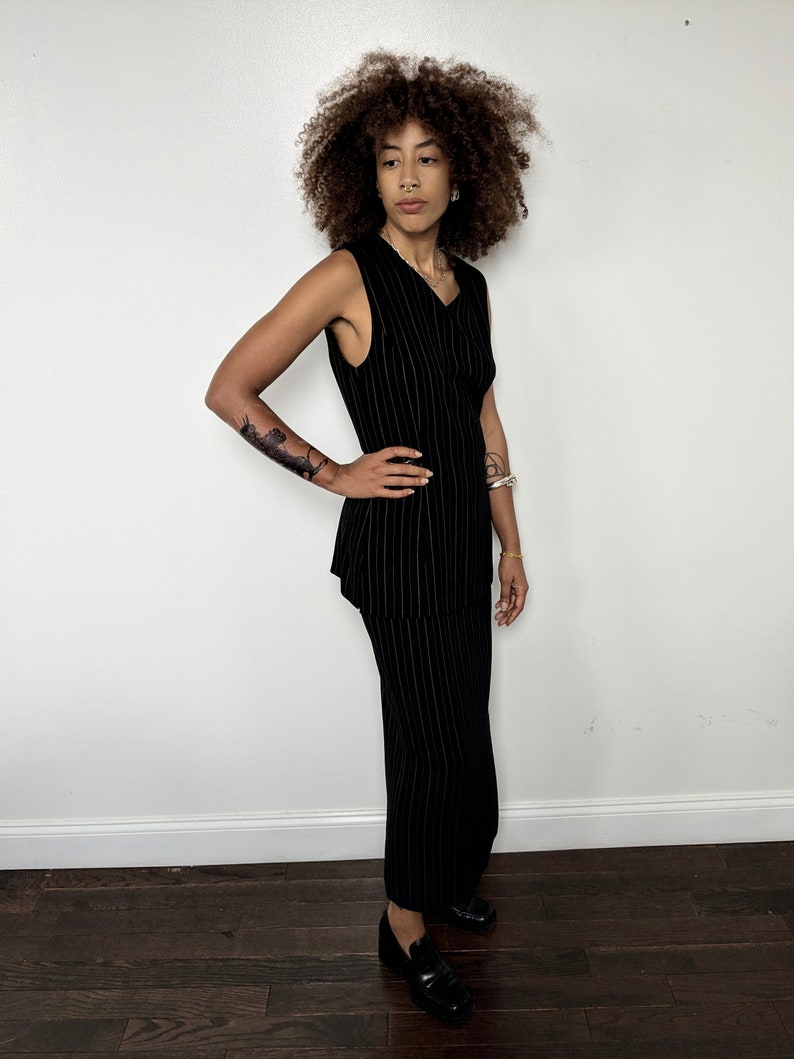 May include: A black sleeveless dress with thin white stripes. The dress has a v-neck and a fitted silhouette. The model is wearing black loafers.