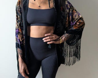 May include: A black velvet kimono featuring a floral pattern in shades of purple, gold, and brown. The garment has long, wide sleeves with black fringe trim along the edges and a solid black velvet border along the front opening and neckline.