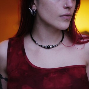 Può includere: Primo piano di una donna che indossa una canotta rossa e una collana nera a choker con perline argentate e nere. La donna ha i capelli rossi e indossa più anelli d'argento all'orecchio sinistro.