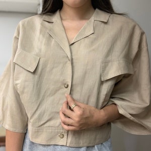 May include: A beige linen shirt with a collared neckline, button closure, and two patch pockets. The shirt has a cropped length and short sleeves.