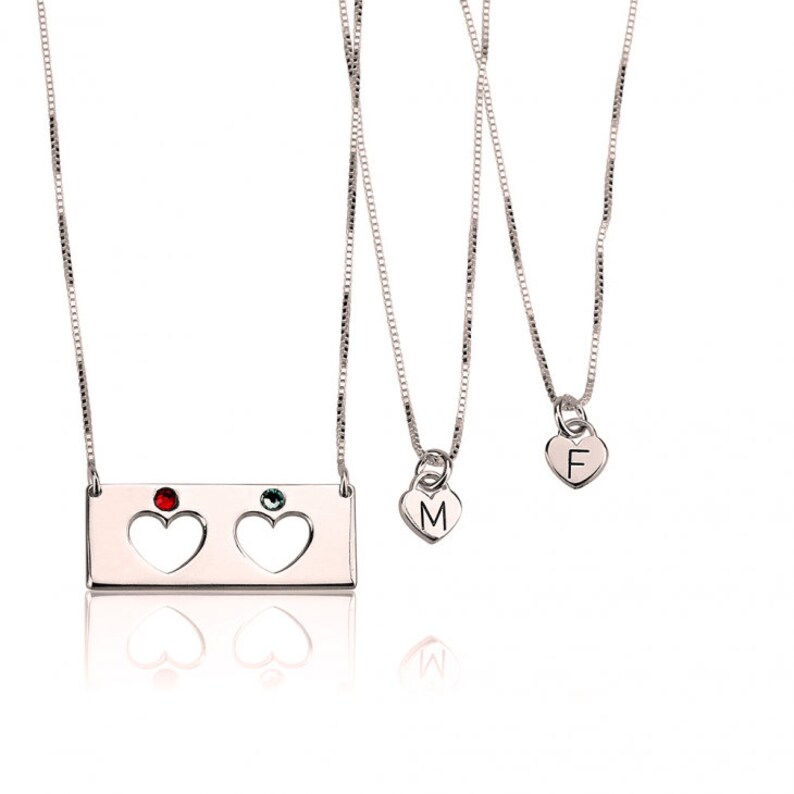 May include: Three silver necklaces with heart pendants. The top necklace has a rectangular charm with two cut-out hearts and red and green gemstones. The middle necklace has a heart pendant with the letter "M" and the bottom necklace has a heart pendant with the letter "F".