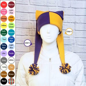 Pode incluir: Um gorro de dois tons com um padrão xadrez roxo e dourado com duas abas longas e pendentes em forma de orelhas. Cada aba termina num pompom multicolorido feito de tiras de tecido roxo e dourado, exibido num manequim.