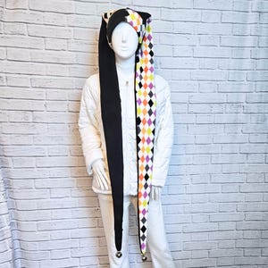 Peut inclure: Un long bonnet de bouffon avec un panneau noir uni d'un côté et un motif à losanges multicolores orange, jaune, violet et noir de l'autre. Le bonnet possède deux longues pointes pendantes, chacune terminée par une petite clochette en métal argenté.