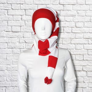 Puede incluir: Un conjunto largo de gorro y bufanda de forro polar suave con un vibrante patrón de rayas horizontales rojas y blancas. La prenda incluye una sección de gorro redondeada que se extiende en una bufanda, terminada con un pequeño pompón rojo.