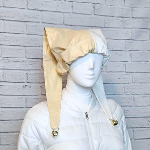 May include: A two-toned jester hat featuring a cream-coloured side and a white side, constructed with a soft, gathered fabric design. The hat includes two long, pointed ear-like flaps that hang down, each finished with a small, shiny gold-toned bell.