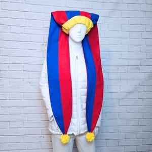 May include: A colorful jester hat with a long, pointed design. The hat features a combination of red, blue, and yellow fabric, with yellow pom-poms at the ends. The hat is displayed on a mannequin.