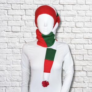 May include: A fleece hat and scarf set featuring a colour-blocked pattern of red, white, and green. The hat has a rounded crown, and the attached scarf features alternating horizontal stripes of red, white, and green, finished with a red pom-pom.