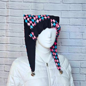 May include: A jester-style hat featuring a black, pink, and blue diamond harlequin pattern. The headpiece has two long, pointed side panels that hang down, each finished with a small metallic bell at the tip. The fabric is black on the underside.