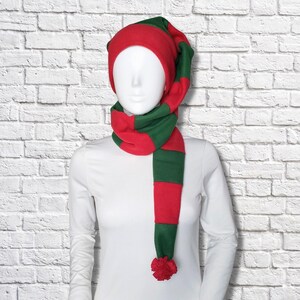 Puede incluir: Un conjunto de gorro y bufanda de forro polar con un patrón de rayas horizontales en rojo y verde oscuro. El gorro tiene una cola larga y puntiaguda que cuelga, y la bufanda está integrada en el diseño, terminando con un pompón rojo.