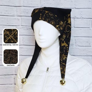 May include: A black jester hat featuring a gold-coloured print of celestial and mystical symbols, including moons, stars, and hands. The hat has two long points, each finished with a small gold-coloured bell at the end, worn on a white mannequin.