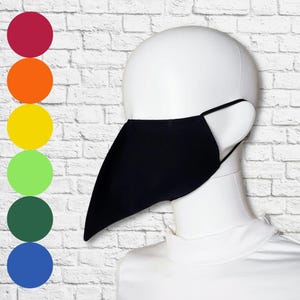 Pode incluir: Uma máscara facial preta com uma projeção pontiaguda em forma de bico que se estende da área do nariz, usada em uma cabeça de manequim branca. À esquerda, uma coluna vertical de seis círculos de cores sólidas exibe vermelho, laranja, amarelo, verde claro, verde escuro e azul.