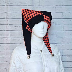May include: A jester hat featuring a black and orange chequered pattern on the pointed ends and solid black fabric on the crown. The two long, dangling points of the hat each feature a small, round silver-coloured metal bell at the very tip.