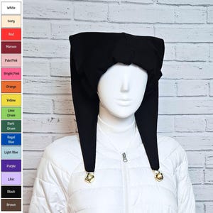 Puede incluir: Un gorro de estilo bufón negro con dos largas solapas laterales puntiagudas que cuelgan, cada una terminada con un pequeño cascabel metálico de color dorado en la punta. El gorro se muestra sobre una cabeza de maniquí blanca con una chaqueta.