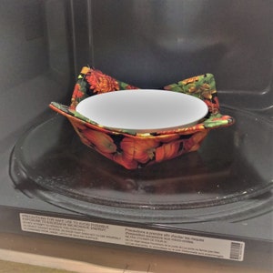 May include: A white ceramic bowl sits inside a colorful microwave bowl cozy. The cozy features a floral pattern with orange, red, and green accents. The cozy is designed to protect hands from hot bowls. The microwave is black.