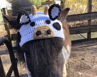 May include: A black and white pony wearing a crocheted cow hat with black and white spots. The hat has a brown nose and white ears.