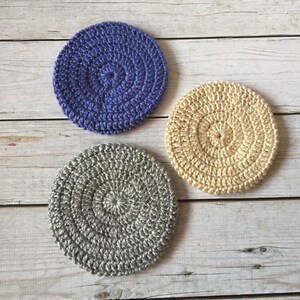 May include: Three crocheted coasters in different colors: purple, beige, and gray. Each coaster has a circular design with a raised center.