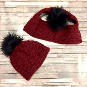 May include: Two crocheted hats with black faux fur pom poms. The hats are burgundy in color. One hat is a slouchy beanie style and the other is a more fitted style.