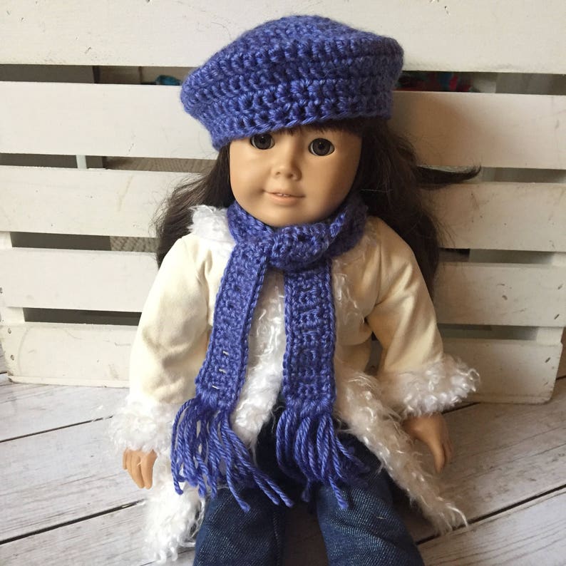 May include: A doll wearing a purple crocheted beret and matching scarf. The doll is wearing a white faux fur jacket and blue jeans.