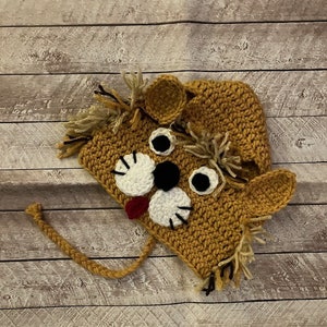May include: A crocheted lion hat with a brown and white face, black eyes, and a red tongue. The hat has a long tail and a mane of brown yarn.