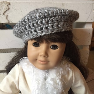 May include: A gray crocheted beret hat for a doll. The hat is made with a simple stitch pattern and has a classic beret shape.