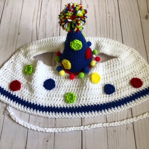 May include: A handmade crochet party hat and bib set. The white bib features colorful polka dots and a blue trim. The blue party hat is adorned with colorful pom-poms and polka dots, topped with a multi-colored pom-pom.
