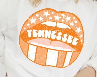 Könnte beinhalten: Weißes Sweatshirt mit einem orangefarbenen Lippen-Grafik-Design im Used-Look, mit dem Wort "Tennessee". Das Lippendesign enthält weiße Sterne und vertikale Streifen, die einen Vintage-Look erzeugen. Das Sweatshirt hat lange Ärmel.