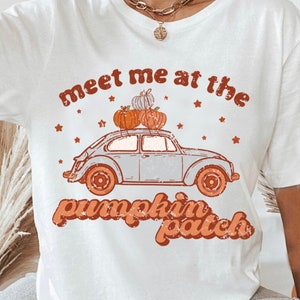 Peut inclure: T-shirt blanc avec un motif rétro. Il présente une voiture bleu clair transportant des citrouilles, avec le texte "Meet me at the pumpkin patch" en orange. Le motif comprend des étoiles oranges et une esthétique vintage.