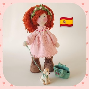 Puede incluir: Muñeca de ganchillo hecha a mano con cabello rojo, vestida con un vestido rosa y botas marrones. La muñeca sostiene una correa sujeta a un perro pequeño de ganchillo. Un bolso verde de ganchillo está al lado del perro. La muñeca tiene una diadema floral verde.