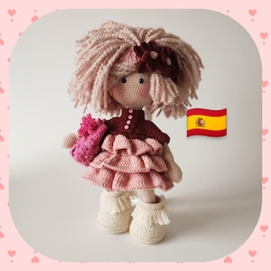 Puede incluir: Una muñeca de crochet con pelo rosa, un jersey burdeos y un vestido rosa con volantes. La muñeca lleva una flor rosa de crochet y lleva botas de color crema con flecos.