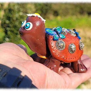 Op de afbeelding: Een bruine schildpadfiguur met blauwe ogen, een witte bloem op zijn kop en een versierd schild met een vlinder, strass en metalen accenten. De schildpad staat op een hand.