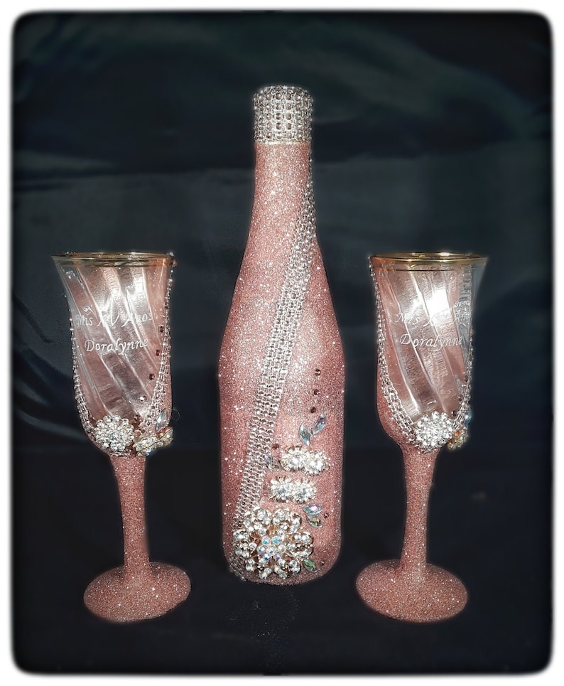 May include: A set of two champagne flutes and a champagne bottle, all decorated with pink glitter and rhinestones. The flutes are engraved with the words "Mrs. & Mr. Doralynn".
