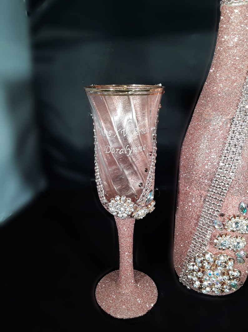 May include: A pink champagne flute with a gold rim and a personalised inscription that reads "Miss XV anos Doralynne". The flute is decorated with glitter and rhinestones.