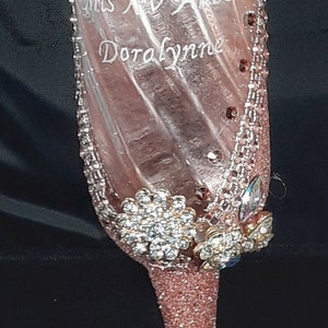 May include: A pink glitter champagne flute with a clear glass body and a gold rim. The flute is decorated with rhinestones and has the text "Mrs. Doralynne" etched on the side.