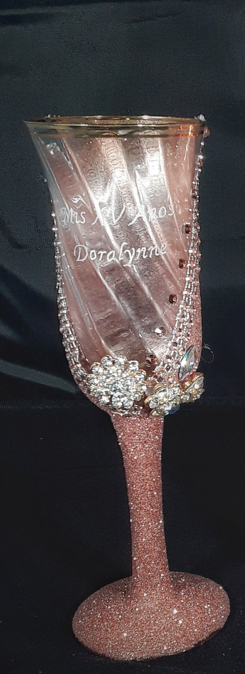 May include: A pink glitter champagne flute with a clear glass body and a gold rim. The flute is decorated with rhinestones and has the text "Mrs. Doralynne" etched on the side.