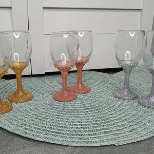 May include: Six wine glasses with glitter accents. Two glasses are gold, two are silver, and two are rose gold.