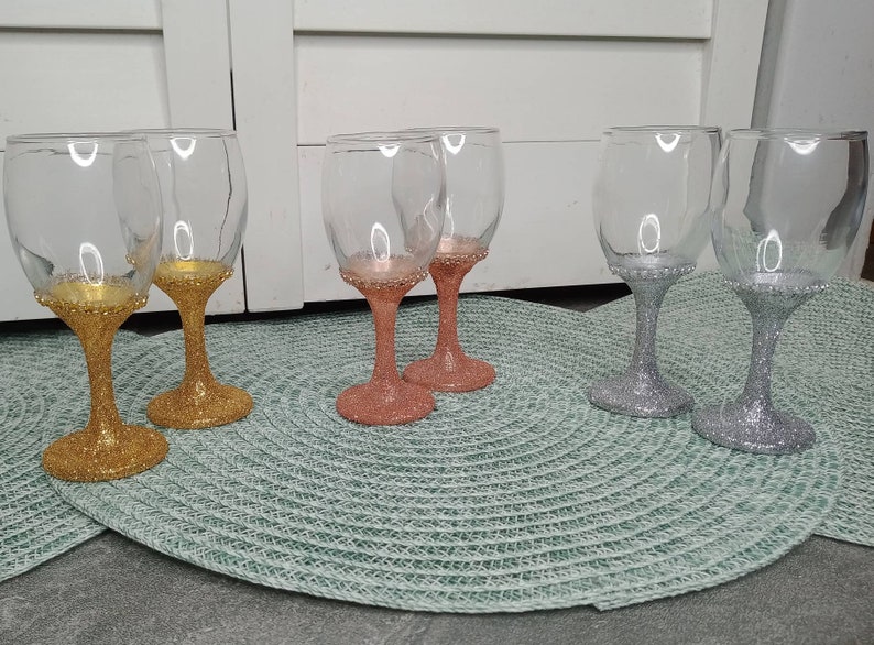 May include: Six wine glasses with glitter accents. Two glasses are gold, two are silver, and two are rose gold.