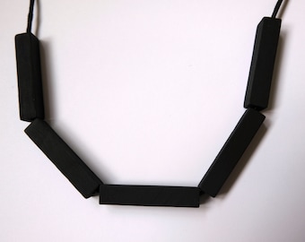 Puede incluir: Un collar negro con cuentas de madera rectangulares. El collar tiene un diseño simple con una cuerda que conecta las cuentas.