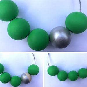 Puede incluir: Un collar hecho de cuentas redondas verdes y plateadas. El collar está hecho de un cordón fino plateado. Las cuentas son grandes y tienen un acabado mate.