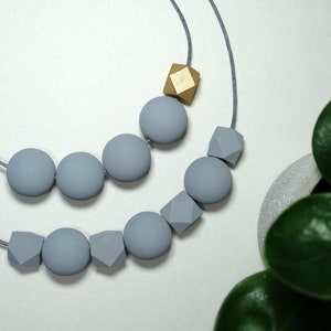 May include: A grey and gold silicone necklace with round and geometric beads. The necklace is made of soft, chewable silicone and is perfect for babies and toddlers.