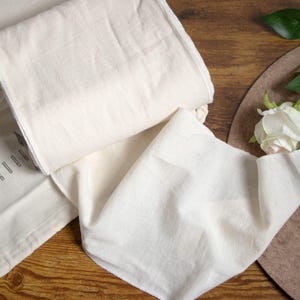 May include: A white cotton fabric runner with a slightly textured surface. The runner is folded in half and laid out on a wooden surface.