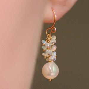 May include: A gold-colored earring with a curved hook. The earring features a cluster of small, iridescent opals and a large, pearly white, teardrop-shaped pearl. The design is delicate and elegant, suitable for special occasions.