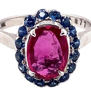 Puede incluir: Un anillo de plata con una gran piedra preciosa ovalada rosa rodeada por un halo de piedras azules más pequeñas.