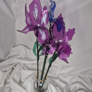 May include: A decorative arrangement of three translucent purple and blue flowers with delicate, thin petals and white centres. The flowers are attached to thin green stems and are displayed in a clear glass bottle filled with small, smooth stones.