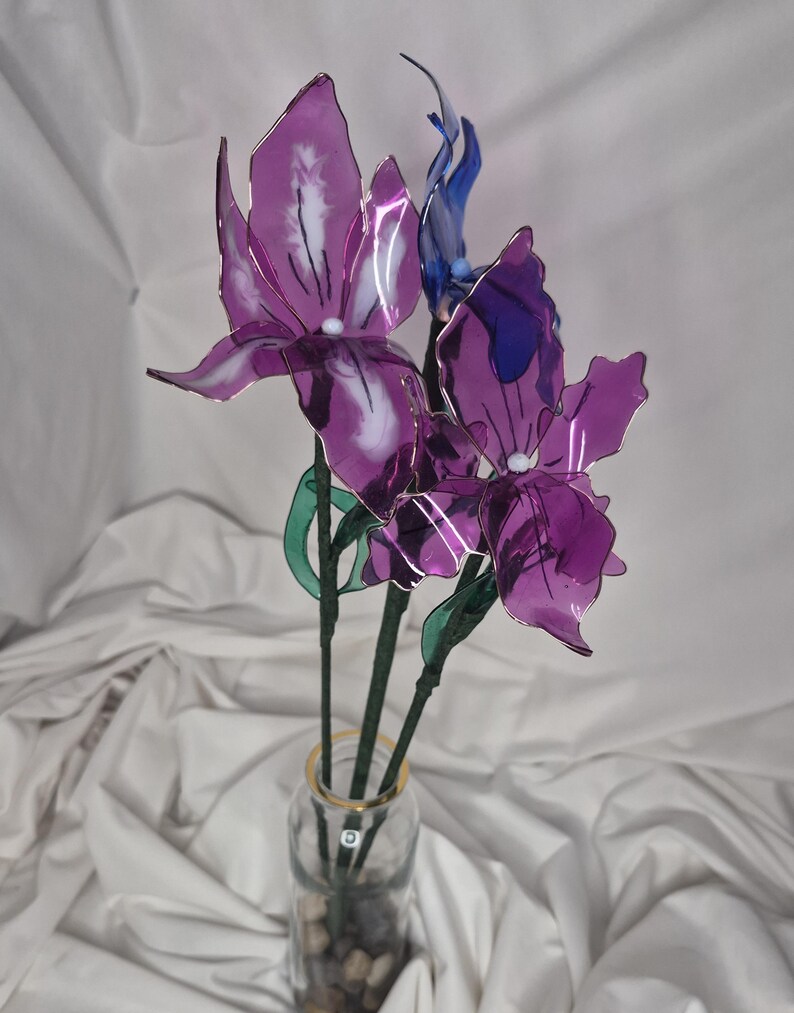 May include: A decorative arrangement of three translucent purple and blue flowers with delicate, thin petals and white centres. The flowers are attached to thin green stems and are displayed in a clear glass bottle filled with small, smooth stones.