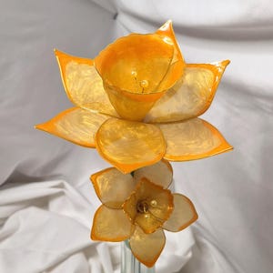 May include: A decorative floral sculpture featuring two daffodil-like flowers with translucent, marbelized orange and yellow petals. The larger top flower and smaller bottom flower both feature central stamens tipped with small, cream-colored beads.