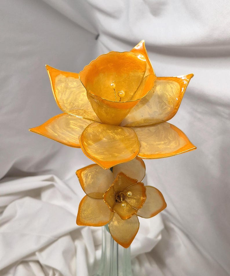 May include: A decorative floral sculpture featuring two daffodil-like flowers with translucent, marbelized orange and yellow petals. The larger top flower and smaller bottom flower both feature central stamens tipped with small, cream-colored beads.
