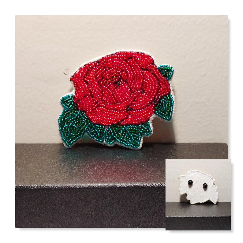 May include: A red beaded rose with green beaded leaves. The rose is attached to a white backing with two black buttons on the back.