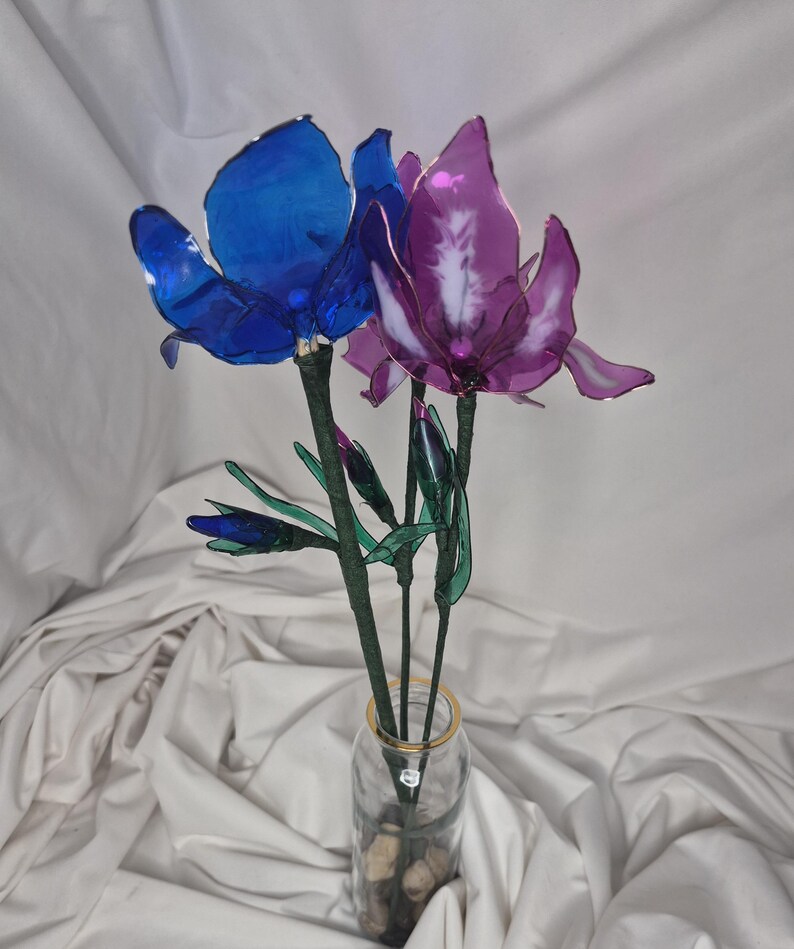 May include: A decorative arrangement of two translucent artificial flowers, one deep blue and one purple, with green stems and small buds. The flowers are displayed in a clear glass vase with a gold rim, containing small stones at the base.