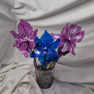 May include: A decorative arrangement of three translucent flowers with thin stems, featuring two purple blossoms and one central blue blossom, each with dark linear details and a small white centre, displayed in a clear glass tumbler.
