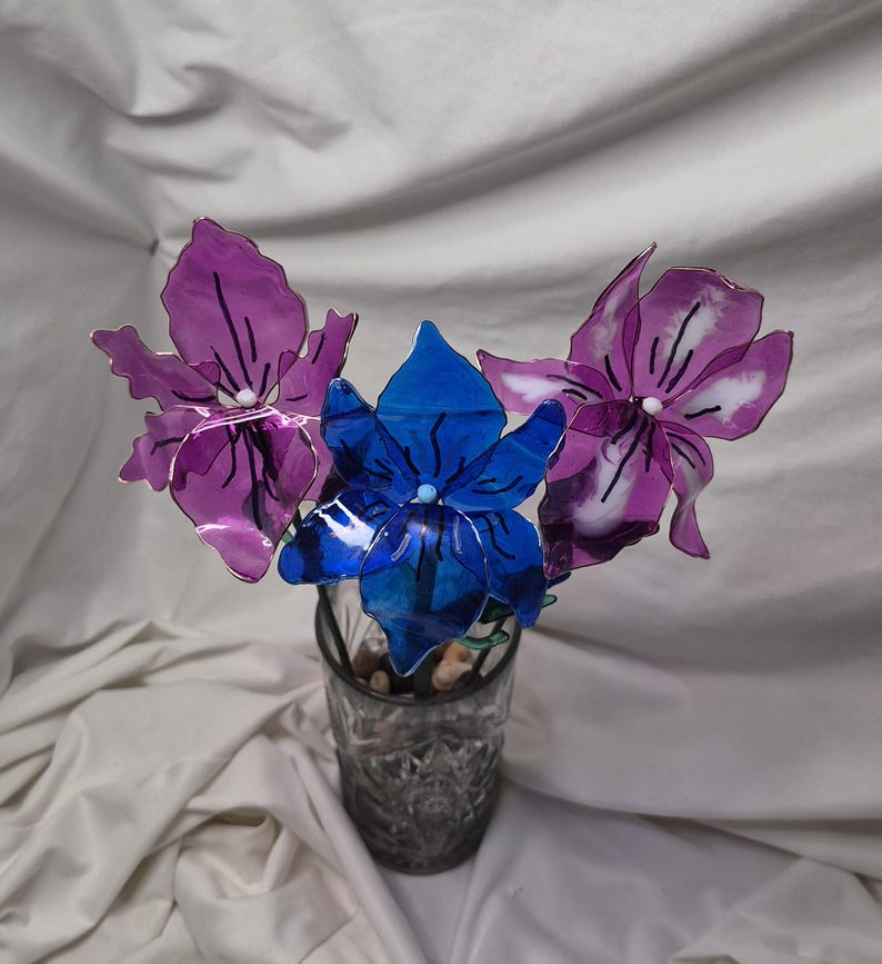 May include: A decorative arrangement of three translucent flowers with thin stems, featuring two purple blossoms and one central blue blossom, each with dark linear details and a small white centre, displayed in a clear glass tumbler.